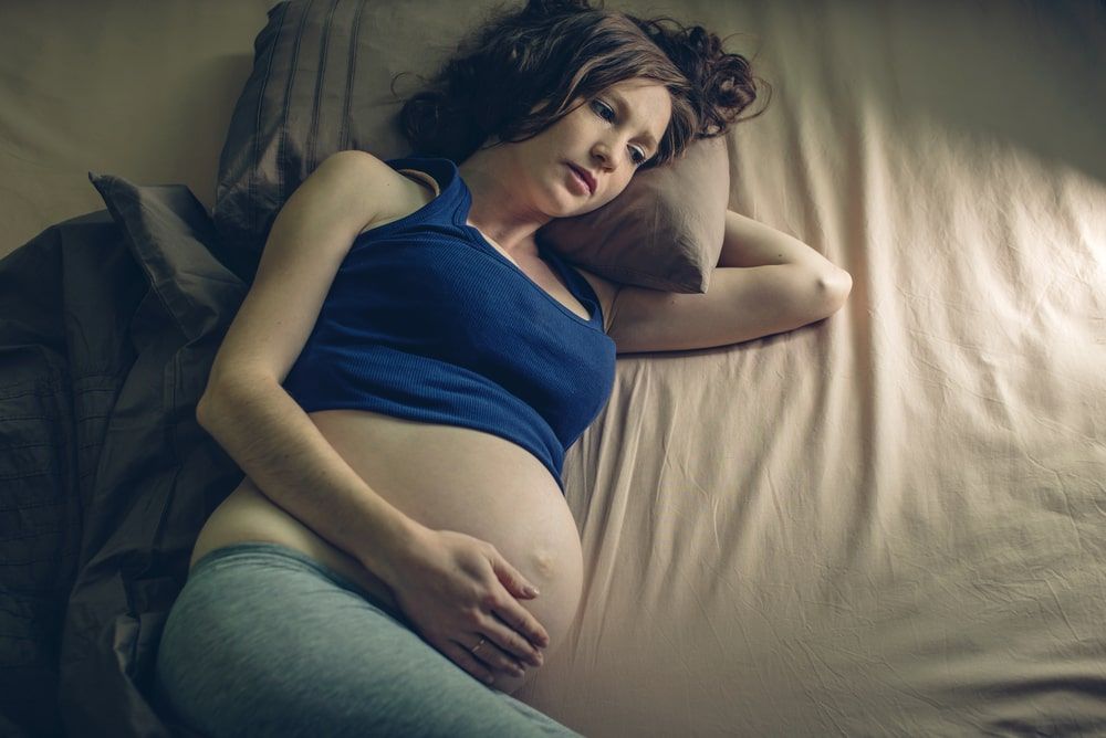 Pregnant woman laying on her side in bed looking forlorn and hopeless with one hand on her pregnant belly. 