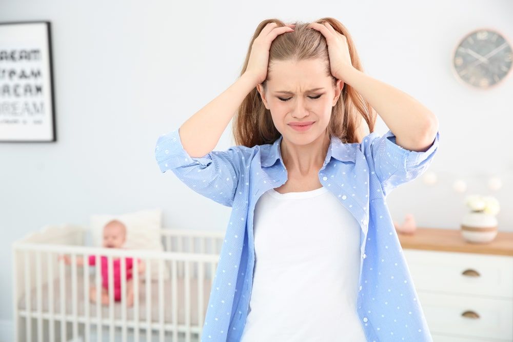 Woman pulling her own hair with an anxious facial expression with a baby in a crib in the background.