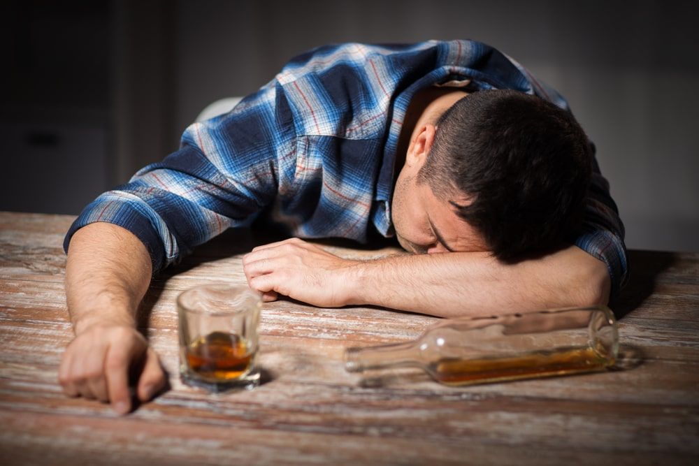 Man passed out with his head on the table and a half-full bottle of alcohol with a cup in front of him